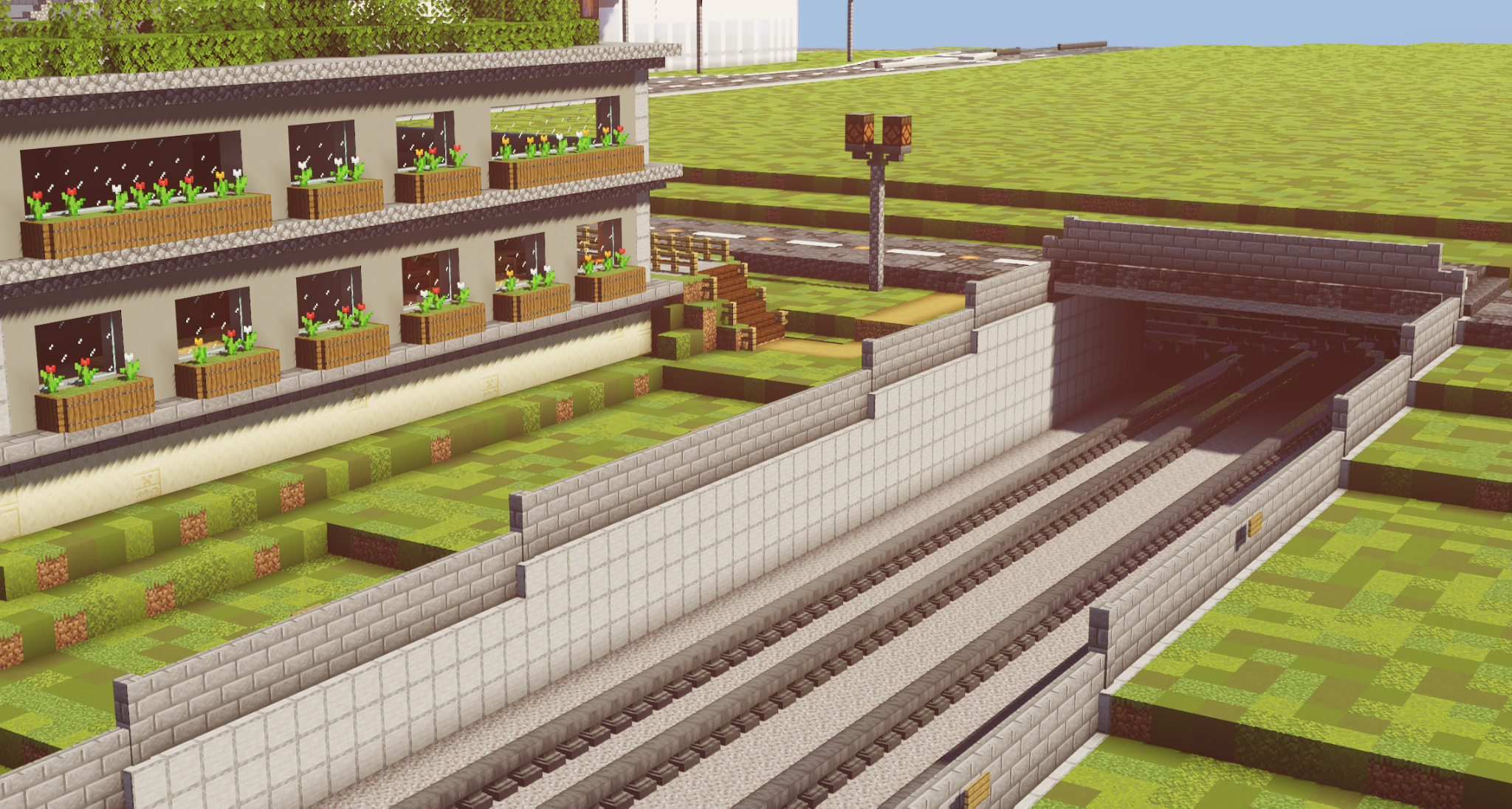 In the foreground is a railroad line with several tracks leading into a dark tunnel on the right-hand side of the picture. The tracks are made of wood and metal. The landscape in which the town is embedded contains green hills, scattered trees and a small field with flowers and plants.